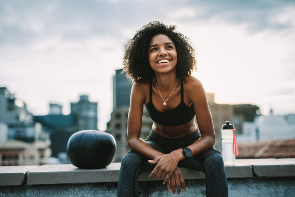 Fitness woman relaxing sitting on rooftop fence looking away. Cheerful female athlete sitting on rooftop fence with a medicine ball and water bottle beside her.