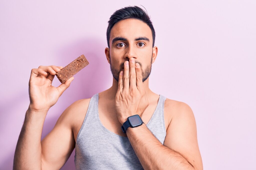 A surprised young athletic man holding a protein bar as he covers his mouth with his hand.