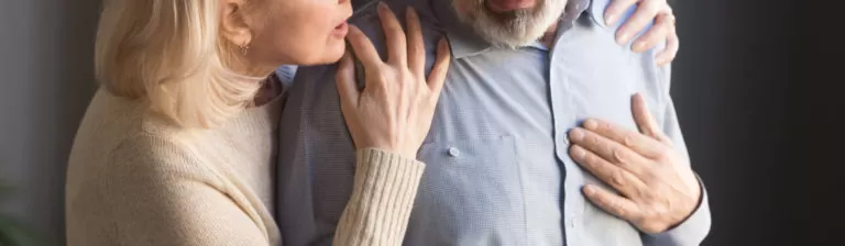 Elderly woman looking concerned as she supports her husband experiencing chest pain, indicating possible heart issues.