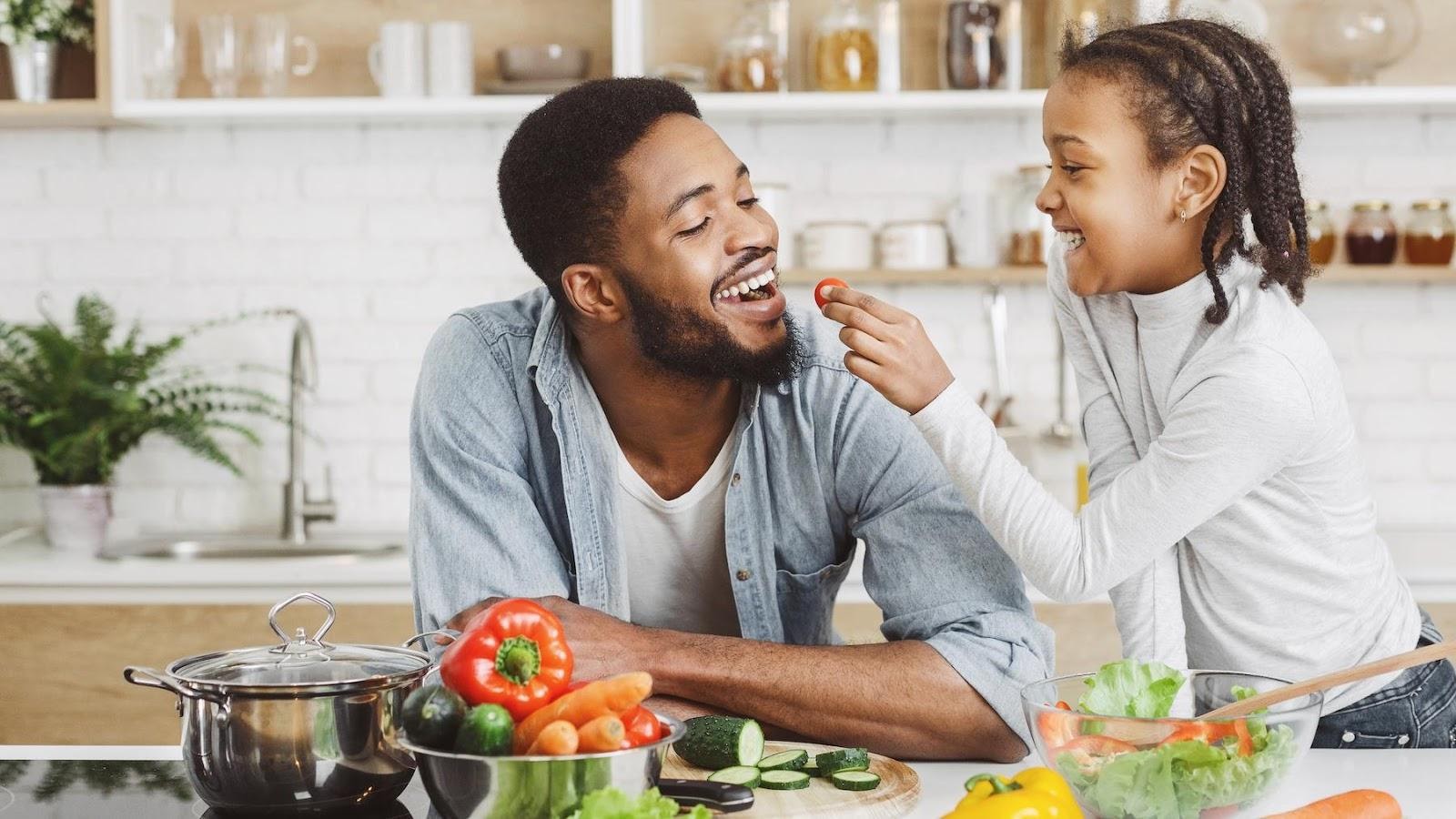 A father and his daughter in the kitchen eating vegetables jovially